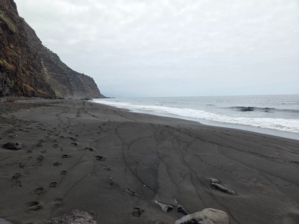 Playa de arena negra casi desierta enmarcada por un frente de acantilados verdes y rojizos.
