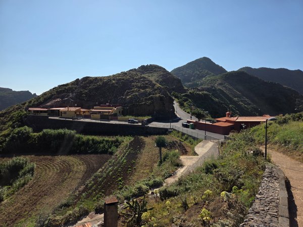 Conjunto de viviendas de Chinamada y la ermita vistos desde el sendero, rodeados de laderas agrícolas.