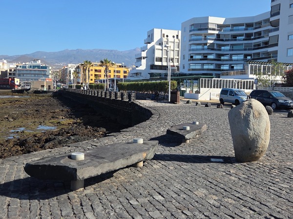 Bancos de piedra y barandillas en el paseo marítimo de El Médano con edificios modernos y marea baja.