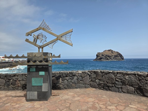 Escultura metálica con forma de pez frente al mar con el Roque de Garachico al fondo.