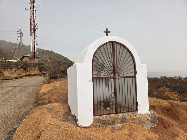 Pequeña capilla blanca con reja y cruz, situada junto a la pista y a las torres de telecomunicaciones.