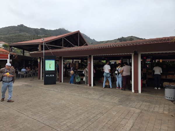 Pasillo de puestos del mercado con las montañas de Tegueste al fondo y pantalla informativa.