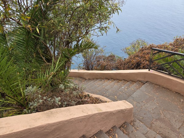 Primer tramo de escalera de piedra con muretes ocres y vegetación costera, descendiendo en zigzag hacia el mar.