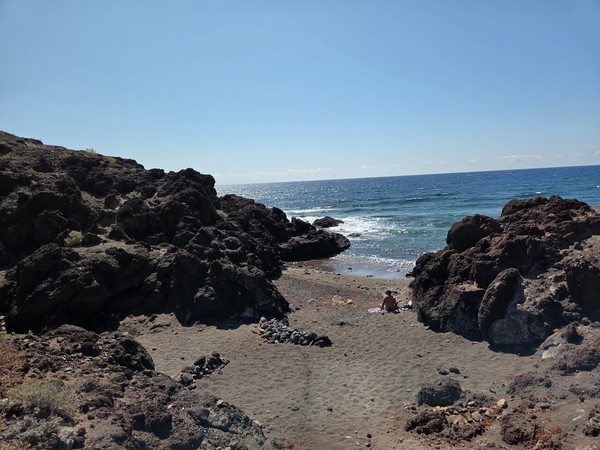 Pequeña cala de arena oscura entre rocas volcánicas, con una persona tomando el sol junto al mar.