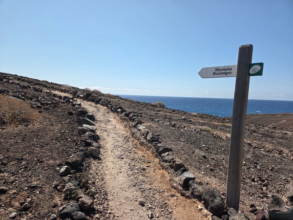 Poste direccional con la indicación Montaña Bocinegro junto al sendero que discurre paralelo al mar.