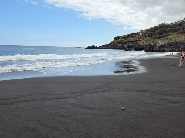 Olas rompiendo suavemente sobre la arena negra de Playa Bollullo con bañistas al fondo
