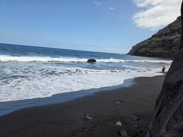 Vista cercana de la orilla de arena negra en Playa El Ancón, con olas rompiendo y un senderista caminando junto al acantilado.