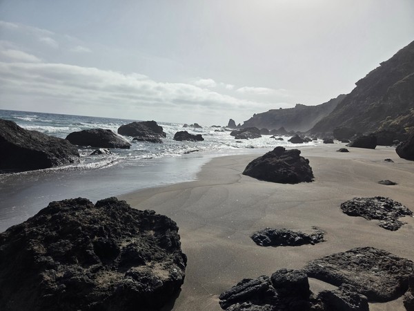 Orilla de arena negra con grandes rocas volcánicas y el Atlántico rompiendo en Playa de Las Breñas.