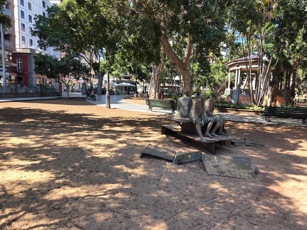 Estatua de un personaje sentado con sombrero sobre pedestal, ubicada junto al quiosco y bajo los ficus.
