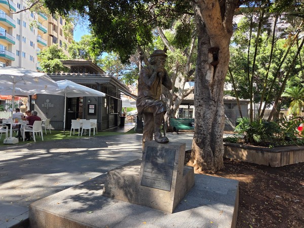 Grupo escultórico de figuras sentadas en un banco dentro de la plaza, con el templete al fondo.