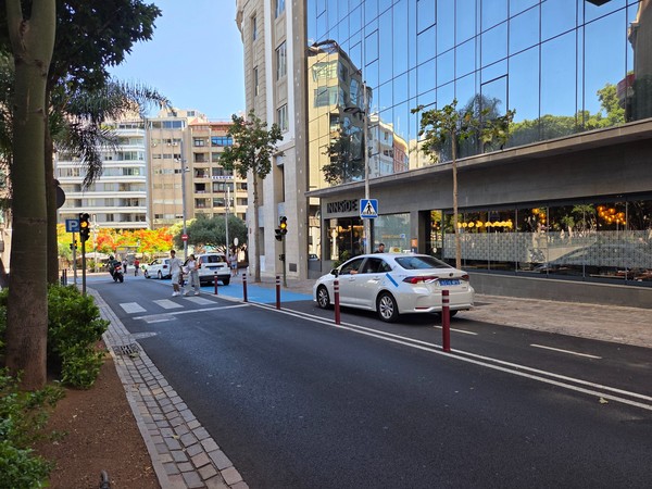 Calle lateral con parada de taxis y paso de peatones junto al límite de la plaza.