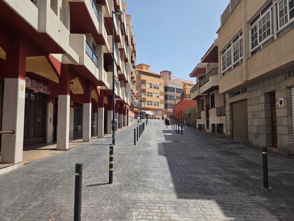 Calle peatonal en ligera pendiente con bolardos y edificios residenciales.