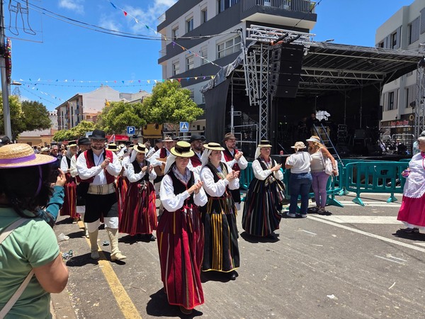 Grupo folclórico avanza aplaudiendo junto al escenario, con vestidos y chalecos de rayas canarias.
