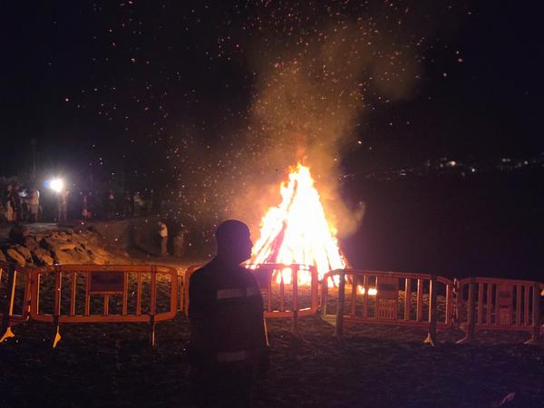 La hoguera arde con llamas altas iluminando la playa y el cielo estrellado.