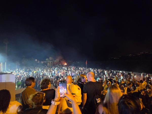 Vista nocturna de la multitud reunida en torno a las hogueras y música en la playa durante San Juan.