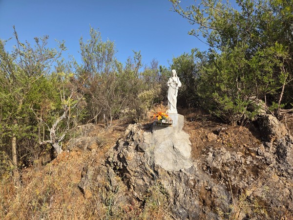 Escultura de Jesús sobre una peana de piedra con flores, en un talud natural.