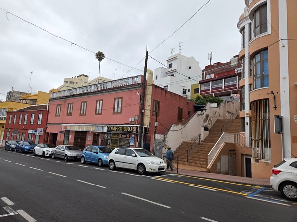 Escaleras y fachadas del centro Tramo de calle en Tacoronte con fachadas color terracota, escaleras urbanas y comercios de proximidad frente a una fila de vehículos estacionados.